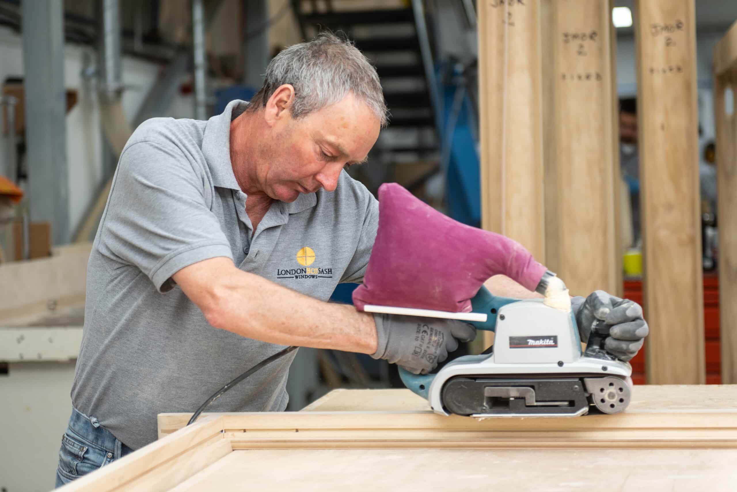 A craftsman sands a timber Accoya sash window in London. Accoya is a sustainable wood alternative that is resistant to rot, decay, and insect damage. This makes it an ideal material for windows in high-traffic areas, such as London. The craftsman is using a makita belt sander to smooth the surface of the window before it is painted or stained.