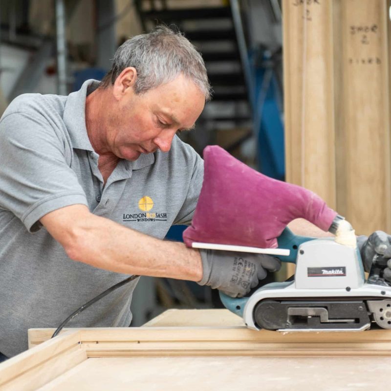 A craftsman sands a timber Accoya sash window in London. Accoya is a sustainable wood alternative that is resistant to rot, decay, and insect damage. This makes it an ideal material for windows in high-traffic areas, such as London. The craftsman is using a makita belt sander to smooth the surface of the window before it is painted or stained.